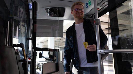 Foto von einem jungen, lächelnden Mann im Bus, der ein VBB-Abo in der Hand hält